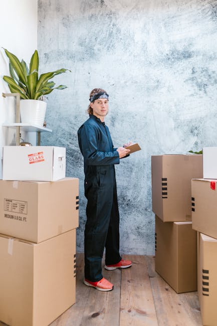 Two men sit inside the open rear of a moving van during a home relocation, with cardboard boxes of various sizes stacked behind them on the van's floor. The man on the left is wearing a beige knit bea