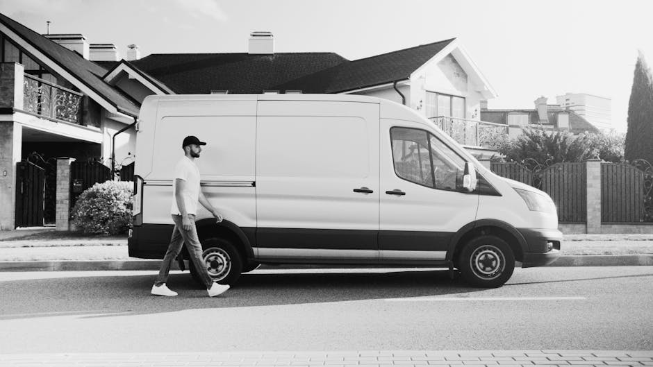 A white moving van labeled 'MOVING COMPANY LOCAL & LONG DISTANCE' is parked on a street in front of a brick building with large arched windows and white walls. The van's side door is closed, and a dri