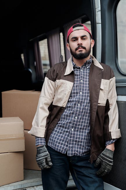 A man with a beard wearing a red cap backwards, a checkered shirt, and a beige and brown jacket stands inside a van during a home relocation process. The van's interior shows a row of windows along th