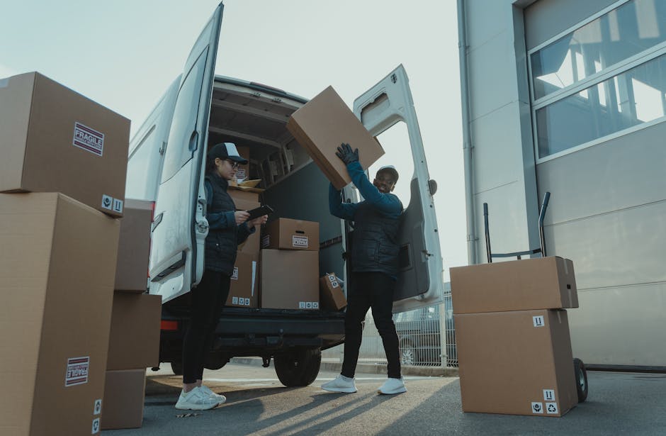 A man with a beard wearing a red cap backwards, a checkered shirt, and a beige and brown jacket stands inside a van during a home relocation process. The van's interior shows a row of windows along th