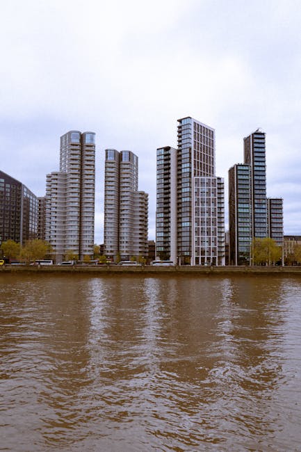 A row of modern high-rise residential and commercial buildings along the riverbank, with large glass windows and concrete facades. The buildings vary in height and architectural style, with some featu