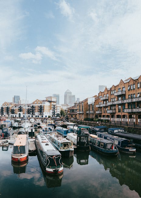 Photograph of a white clock tower situated on a concrete quay by a body of water, with a cluster of mid-rise brick and glass buildings in the background under a blue sky with some wispy clouds. The im