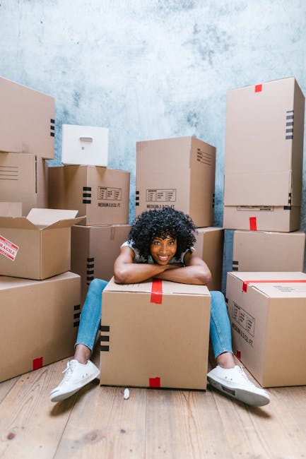 A woman with long, curly hair wearing a grey sweatshirt is inside a property, holding two cardboard boxes securely. One box, marked with the word 'TOYS' in large handwriting, is placed on top of a lar