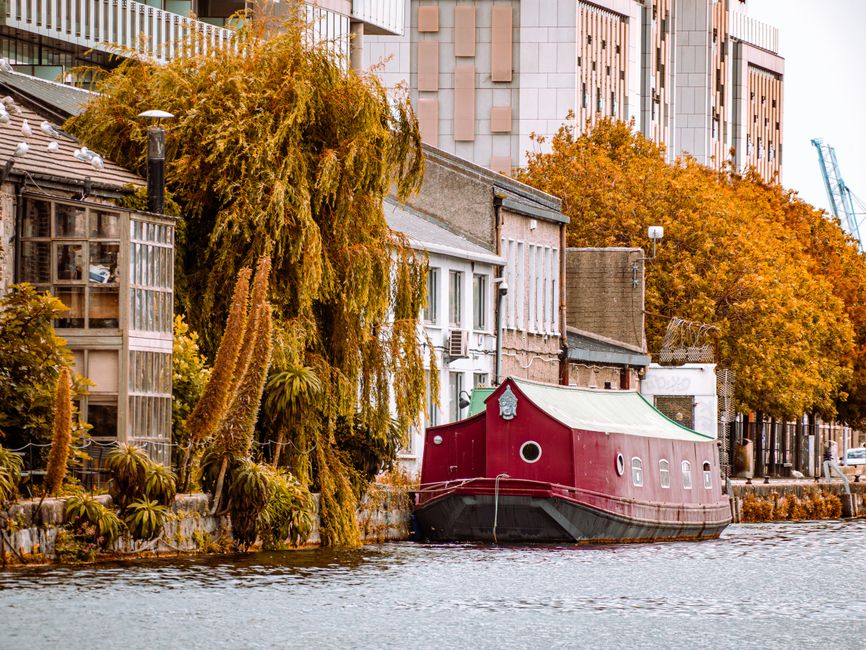 A narrowboat painted in dark red with a curved, light-colored roof is moored along the edge of a canal, adjacent to a row of residential buildings that include a modern multi-storey structure with lar