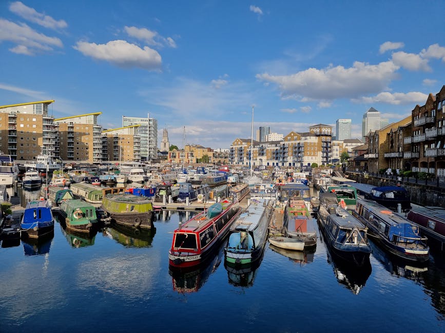 A narrowboat painted in dark red with a curved, light-colored roof is moored along the edge of a canal, adjacent to a row of residential buildings that include a modern multi-storey structure with lar