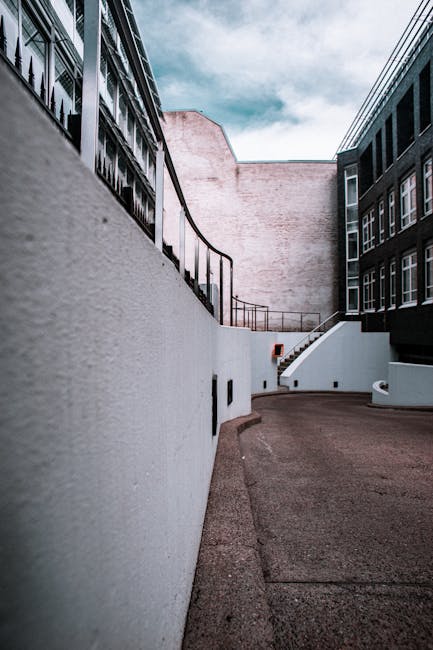 A narrow urban alleyway designed for home relocation activity, featuring a sloped concrete pathway bordered by white painted walls on the left and dark-colored modern buildings with multiple windows on the right. The pathway leads to a set of concrete stairs with metal railings visible in the background, which may facilitate access to a building or exterior level. Overhead, a partly cloudy sky is visible, providing natural light that illuminates the area evenly. The scene implies a confined access point suitable for furniture transport and loading processes, with no visible vehicles or moving equipment. This environment is associated with property removals and moving services, such as those provided by Man and Van Wapping, focusing on narrow or restricted access properties, as highlighted in the page titled 'Wapping High Street removals for narrow access properties, WAPPING'.