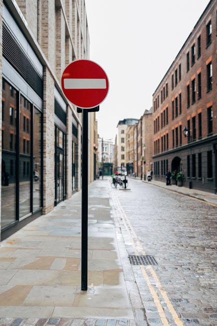 A close-up view of a red and white no-entry traffic sign mounted on a black metal pole situated on a narrow, cobbled street in Wapping, London. The street is lined with multi-storey brick and stone buildings featuring large windows, some with reflective glass. In the distance, a few bicycles and pedestrians are visible, along with additional buildings that create an urban environment typical of house removals and relocation settings. The pavement shows a mix of concrete slabs and cobbles, with a drainage grate near the sign. Bright, natural daylight illuminates the scene, emphasizing the architecture and street details, consistent with a context involving moving or transport logistics in residential or commercial properties, as managed by services such as Man and Van Wapping.