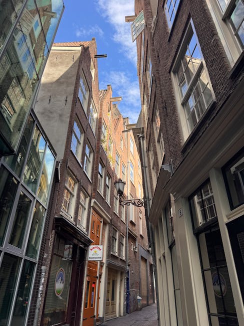 A narrow urban alleyway designed for home relocation activity, featuring a sloped concrete pathway bordered by white painted walls on the left and dark-colored modern buildings with multiple windows on the right. The pathway leads to a set of concrete stairs with metal railings visible in the background, which may facilitate access to a building or exterior level. Overhead, a partly cloudy sky is visible, providing natural light that illuminates the area evenly. The scene implies a confined access point suitable for furniture transport and loading processes, with no visible vehicles or moving equipment. This environment is associated with property removals and moving services, such as those provided by Man and Van Wapping, focusing on narrow or restricted access properties, as highlighted in the page titled 'Wapping High Street removals for narrow access properties, WAPPING'.